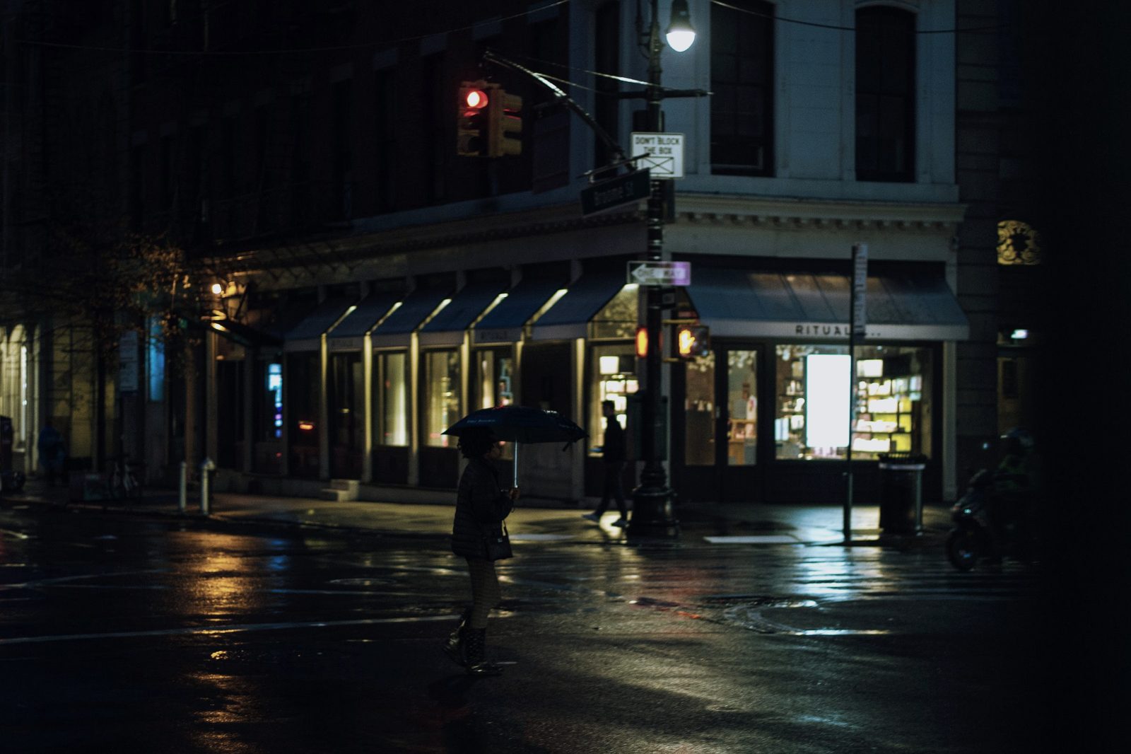 A wet street corner at night, one figure crossing under an umbrella.